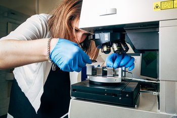 Das Bild zeigt die Forscherin, wie sie mit Handschuhen ein kleines Glasstück unter dem Mikroskop untersucht. 