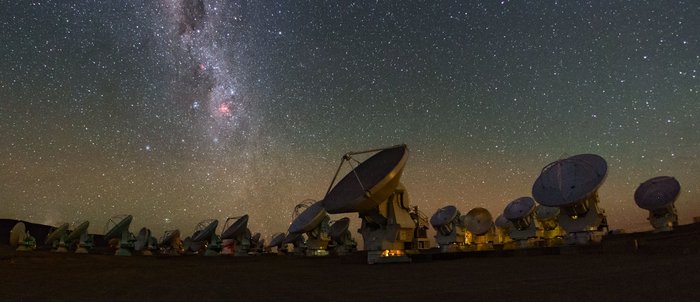 Die Radioteleskopanlage ALMA Parabolantennen in der Wüste unter einem Sternenhimmel, an dem sich die Milchstraße abzeichnet.