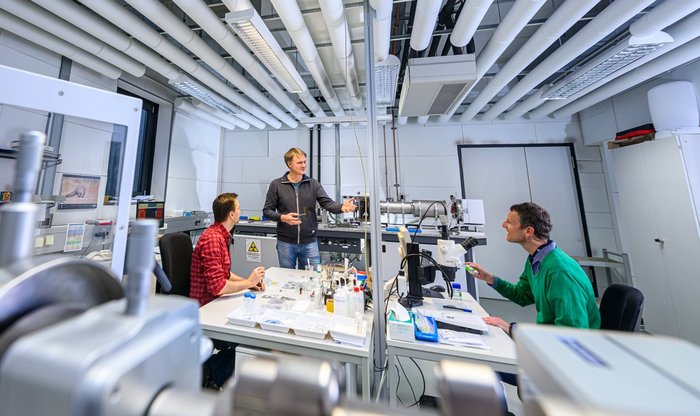Drei Wissenschaftler diskutieren miteinander an Tischen mit Forschungsinstrumenten in einem Labor.