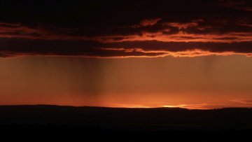 Eine untergehende Sonne am Horizont färbt eine Landschaft und Regenwolken rot