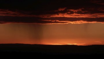 Eine untergehende Sonne am Horizont färbt eine Landschaft und Regenwolken rot
