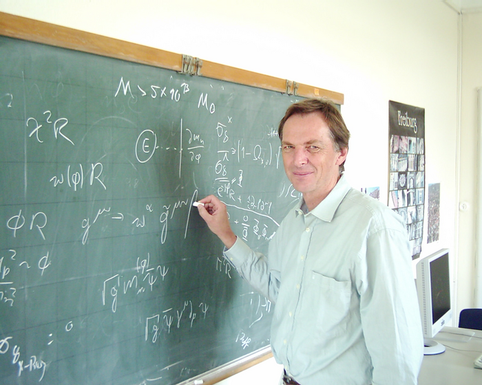 Christof Wetterich Das Bild zeigt den Forscher rechts von einer Tafel, auf die er gerade mit Kreide schreibt.