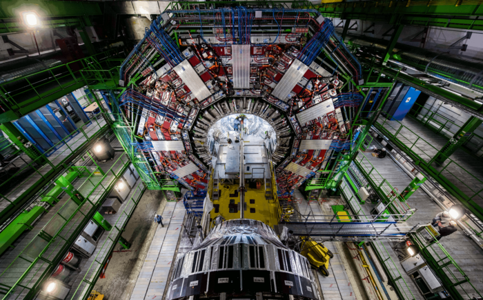 CMS-Detektor Das Bild zeigt eine etwa zwanzig Meter hohe, ringförmige Maschine. In der Mitte des Rings stehen einige Techniker.