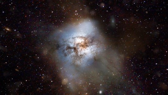 Hell leuchtende, mit einem Schimmer überzogene Galaxie umgeben von dunklem Hintergrund mit einzelnen hellen Punkten