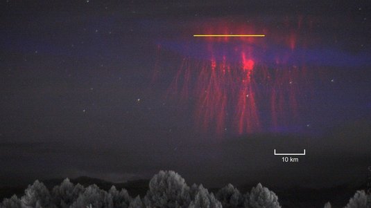 Baumkronen am unteren Rand, darüber ein rotes Leuchten. Im oberen Bereich des Leuchtens ist eine gelbe horizontale Linie, die den Spalt des Spektrografen, mit dem das Team die Messungen gemacht hat, auf das Bild projiziert.  