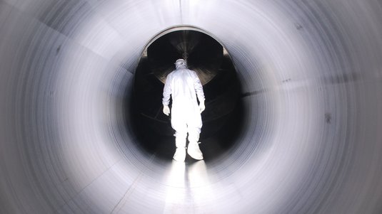 Ein Mann in einem weißen Schutzanzug läuft durch eine weiße Röhre auf ein dunkles Ende des Tunnels zu.