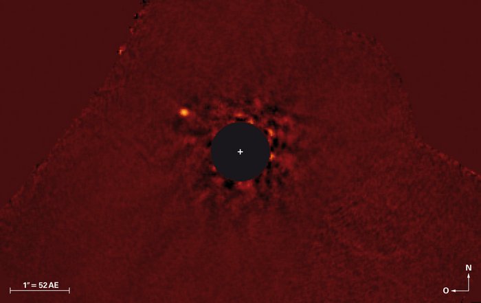 Bearbeitete Aufnahme, der Hintergrund ist tiefrot, in der Mitte ist ein schwarzer, runder Fleck zu sehen. An dieser Stelle befindet sich der Stern, dessen Licht herausgefiltert wurde. Links über ihm leuchtet schwach der Planet als weißer Punkt.