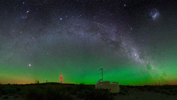 Eine Nachtaufnahme zeigt einen klaren Sternenhimmel mit einem Leuchten über dem Horizont. Im Vordergrund ist einer der Wassertanks des Pierre-Auger-Observatoriums, im Hintergrund eines der Fluoreszenzteleskope.