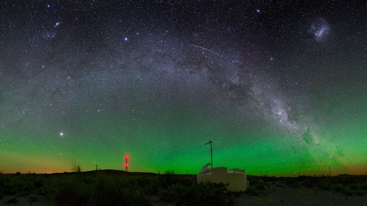 Eine Nachtaufnahme zeigt einen klaren Sternenhimmel mit einem Leuchten über dem Horizont. Im Vordergrund ist einer der Wassertanks des Pierre-Auger-Observatoriums, im Hintergrund eines der Fluoreszenzteleskope.