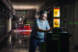 Ein Wissenschaftler steht in einem dunklen Labor. Vor ihm steht auf einem Sockel eine beleuchtete Säule, in der sich Organe in einer Flüssigkeit befinden. Auf dieser Säule liegt das Kreuz, das zwei sich überschneidende Laserstrahlen bilden. 