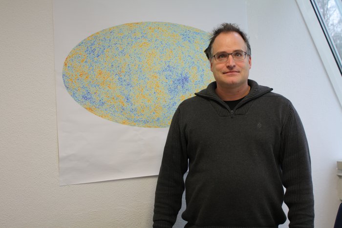 Torsten Enßlin vom Max-Planck-Institut für Astrophysik in Garching vor einer Wand mit einem Poster, dass eine schematische Darstellung der Hintergrundstrahlung zeigt.