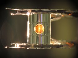 Transparenter Zylinder, der zwischen zwei Platten eingespannt ist. Im Inneren des Zylinders befindet sich ein goldfarbener, kugelförmiger Hohlraum.