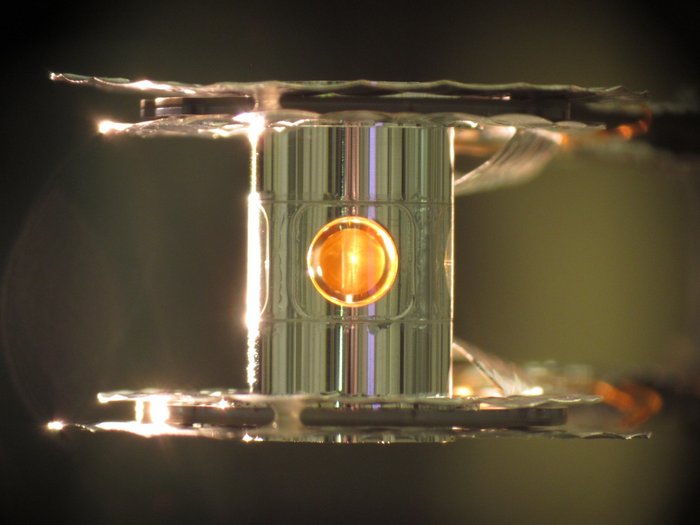 Transparenter Zylinder, der zwischen zwei Platten eingespannt ist. Im Inneren des Zylinders befindet sich ein goldfarbener, kugelförmiger Hohlraum.