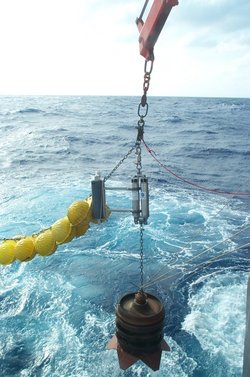 Foto. Blick von einem Schiff aufs Meer. An einem Kran des Schiffs hängt eine Messsonde mit ein paar daran befestigten Bojen. Die Sonde wird gerade ins Wasser gelassen.