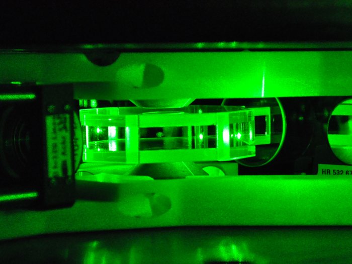 Eine flache, kantige Glaszelle mit gefangenen Atomen, hell erleuchtet von grünem Laserlicht.