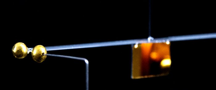 Gravitationswaage Eine Metallstange hängt an einem Faden, an einer Seite befinden sich mehrere goldene Kügelchen, in der Mitte hängt eine Goldfolie