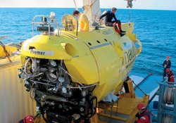 Ein kleines gelbes U-Boot auf seinem Trägerschiff. Zwei Personen befinden sich auf dem U-Boot.