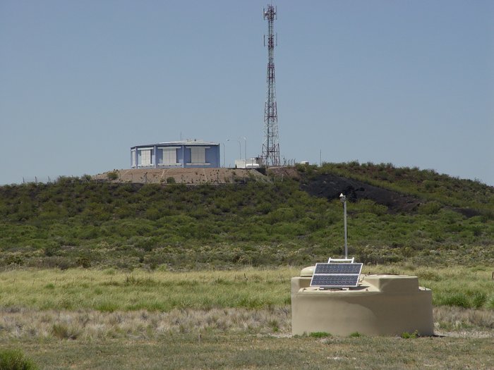 Weißer Zylinder in einer spärlich bewachsenen Steppe. Im Hintergrund auf einer Anhöhe ein Gebäude mit einem antennenähnlichen Mast.