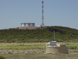 Weißer Zylinder in einer spärlich bewachsenen Steppe. Im Hintergrund auf einer Anhöhe ein Gebäude mit einem antennenähnlichen Mast.