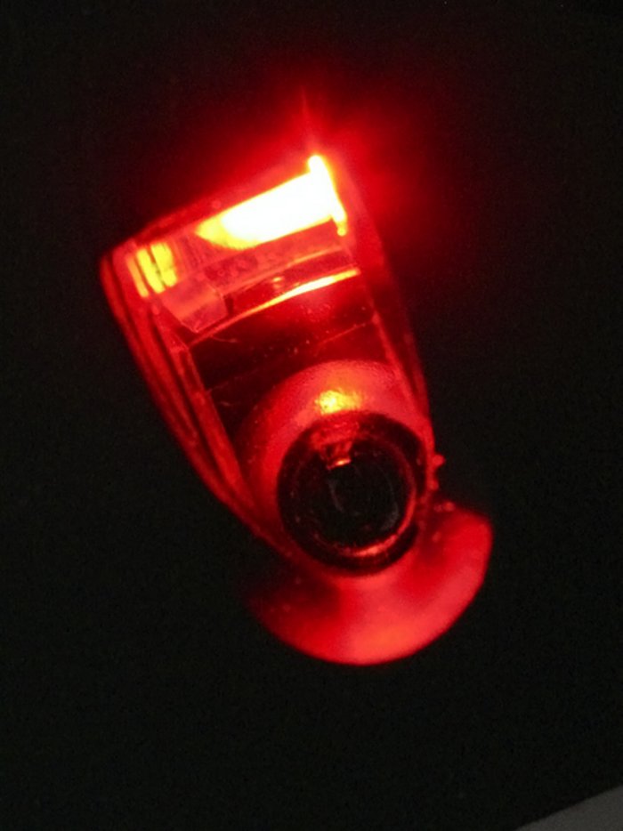 Nahaufnahme der Lichtquelle-Linse-Kombination Auf schwarzem Hintergrund ist vorne der Kreis der Linse zu sehen, klares glänzendes Plastik über einem schwarzen Loch, von rotem Licht umrandet. Daneben eine trapezförmige Struktur, von rotem Licht durchflutet, teils mit gelblichem Licht an den Kanten.
