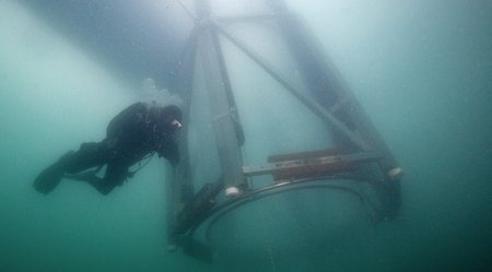 Ein Taucher im Taucheranzug schwimmt unter Wasser. Vor ihm sieht man ein zylinderförmiges Netz.
