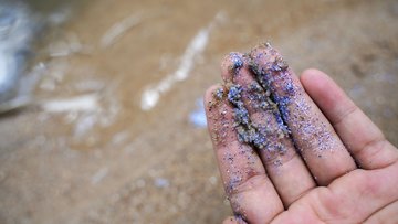 Eine Hand hält viele kleine und bunte Plastikpartikel. Im Hintergrund sieht man den Strand.