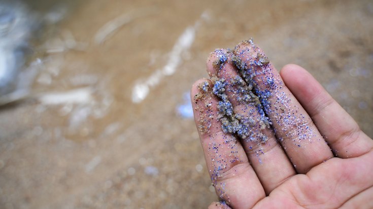 Eine Hand hält viele kleine und bunte Plastikpartikel. Im Hintergrund sieht man den Strand.