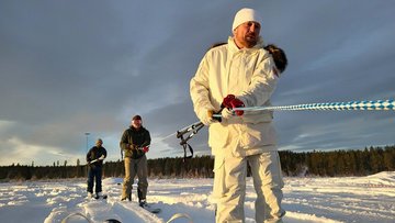 Mehrere Menschen laufen hintereinander Ski in einer Winterlandschaft; die Person ganz vorn trägt weiße Kleidung.