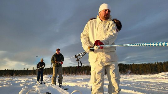 Mehrere Menschen laufen hintereinander Ski in einer Winterlandschaft; die Person ganz vorn trägt weiße Kleidung.