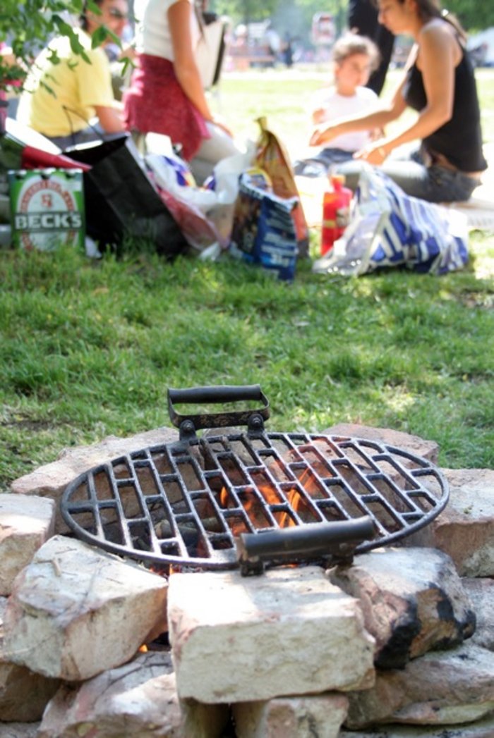 Ein Feuer lodert auf einem Holzkohlegrill, im Hintergrund sind verschwommen Menschen zu sehen.