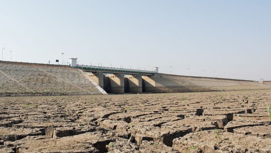 Ausgestrockneter Stausee in Indien | Bildquelle: lakshmiprasad S/iStock Ausgetrocknete Erdoberfläche mit tiefen Rissen, im Hintergrund eine Staumauer