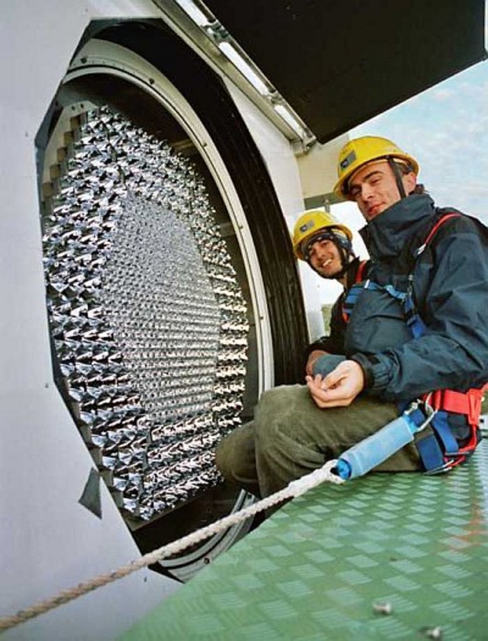 Zwei Physiker beim Einbau der Kamera Auf einer grünen Metallplatte sitzen zwei mit einem Seil gesicherte Personen. Vor ihnen ein geöffneter Bereich des Teleskops, in dem zahlreiche wabenartige silbrig glänzende Strukturen zu sehen sind.