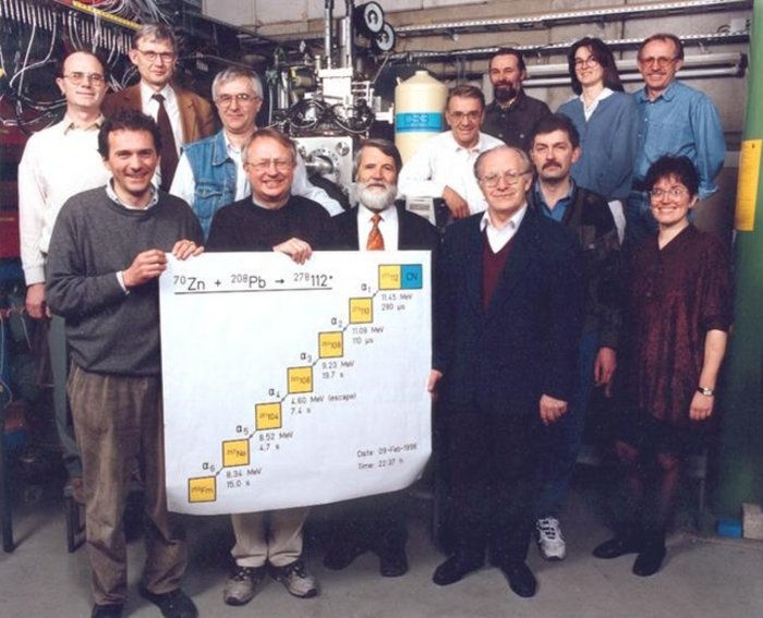 Auf dem Foto ist eine Gruppe von 13 Menschen vor technischen Geräten zu sehen. Vorne halten mehrere Personen ein großes Plakat hoch, auf dem die Zerfallskette des Elements 112 zu erkennen ist.