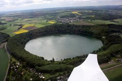 Luftaufnahme eines fast kreisrunden Sees in einer Landschaft aus Feldern, Wiesen und Wäldern.