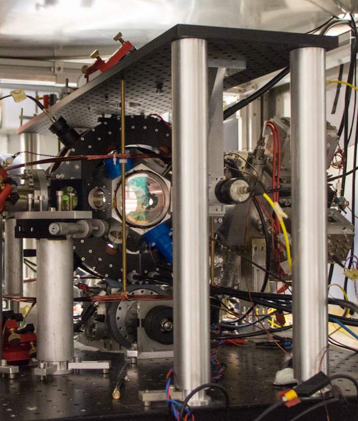 Atominterferometer Auf einem Tisch mit einer schwarzen Oberfläche, auf der sich zahlreiche Löcher für otische Aubauten befindet steht das Experiment. Im Zentrum ist eine reflektierende Linse zusehen, daumherum Kabel und technische Geräte.