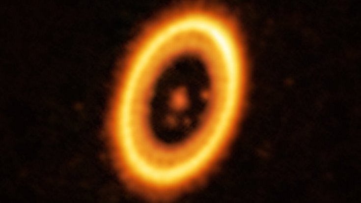 Ein Planet und sein Trojaner im System PDS 70 | Bildquelle: ALMA (ESO/NAOJ/NRAO) /Balsalobre-Ruza et al. Leuchtendes Zentrum mit umgebendem Leuchtring und zwei weiter innen liegenden rötlich leuchtenden Punkten, die gleich weit vom Zentrum des Rings entfernt sind.
