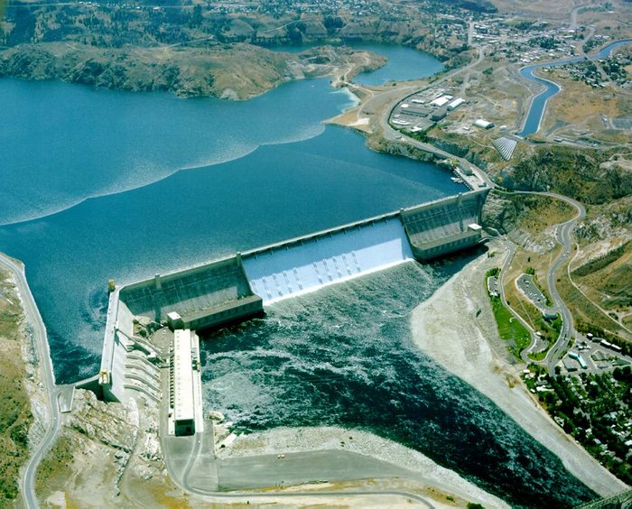 Wasserkraftwerk „Grand Coulee Dam“ in den Vereinigten Staaten Foto aus Vogelperspektive: Eine schräg gestellte Betonmauer trennt einen großen Stausee von einem Flussbett, in dem das Wasser wieder abläuft. Die Mauer ist an der linken Seite abgeknickt, zu Ihrem Fuß liegt das Turbinenhaus. An den Ufern Straßen und Bebauung.