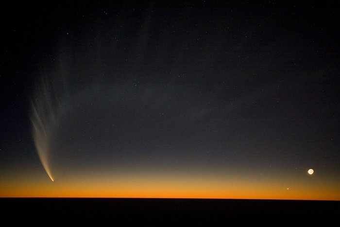 Komet McNaught Vor dunkelblauem Hintergrund sind etliche helle Punkte zu sehen. Links gehen von einem besonders hellen Punkt weiße fächerförmige Strukturen aus, die sich im großen Bogen bis an den rechten Bildrand erstrecken. Rechts unten ein heller Lichtfleck.