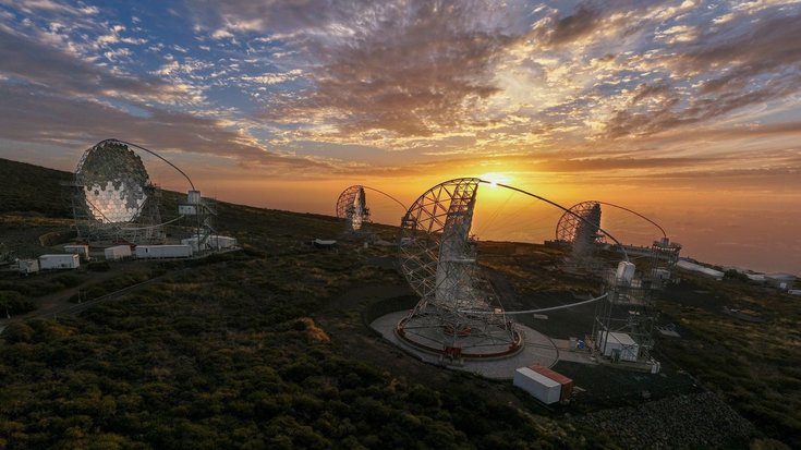 Vier Antennenanlagen mit riesigen gebogenen Spiegeln auf einem mit Bäumen beachsenen Berg, im Hintergrund geht die Sonne unter