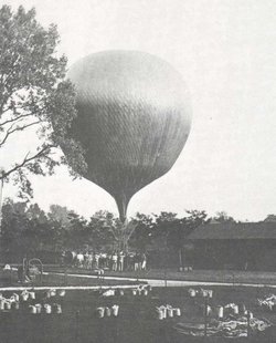 Schwarzweiss-Fotographie: Grosser Heissluftballon steigt auf in einer Gartenlandschaft, viele Besucher schauen zu.
