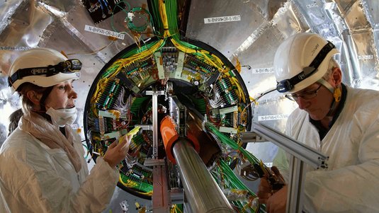 Das Bild zeigt eine lange Metallröhre, die durch die Mitte eines kreisförmigen Experiments läuft. Rechts davon stehen zwei Männer.