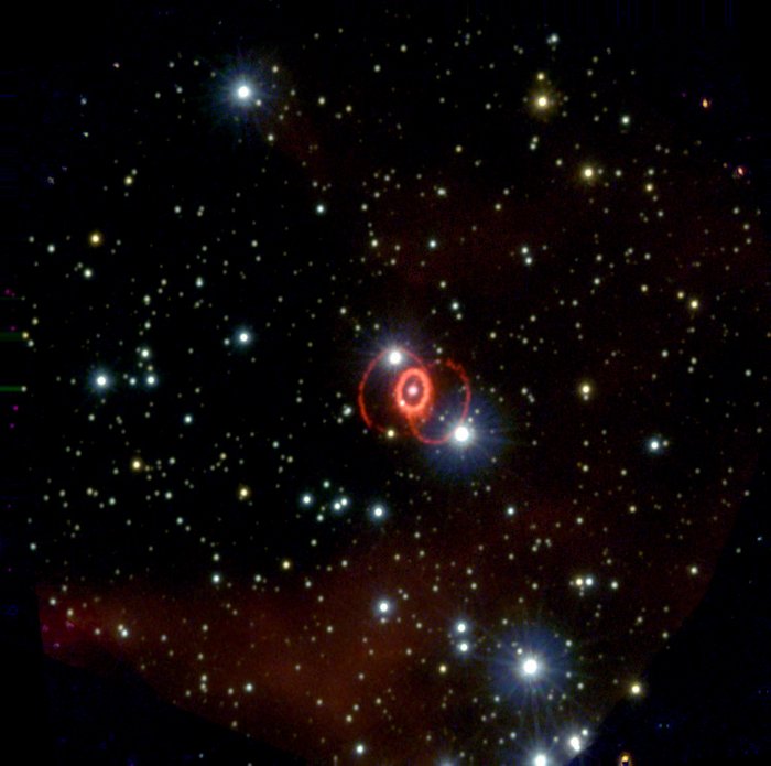 Vor dunklem Hintergrund einige Sterne und rote Wolken; in der Mitte ein Stern, umgeben von einem hellen Ring, an den sich zwei rötlich schimmernde Bögen wie Henkel anschließen.