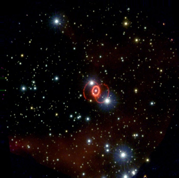 Supernova 1987A Vor dunklem Hintergrund einige Sterne und rote Wolken; in der Mitte ein Stern, umgeben von einem hellen Ring, an den sich zwei rötlich schimmernde Bögen wie Henkel anschließen.