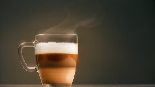 Das Bild zeigt ein Glas mit Latte macchiato. Ganz oben ist Milchschaum, darunter eine Schicht dunkler Espresso, gefolgt von heller werdenden Schichten aus Milch und Espresso.