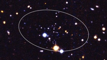 Eine eingekreiste Ansammlung schwach leuchtender Sterne im Zentrum und einige weitere Galaxien und Sterne am Rand vor dunklem Hintergrund
