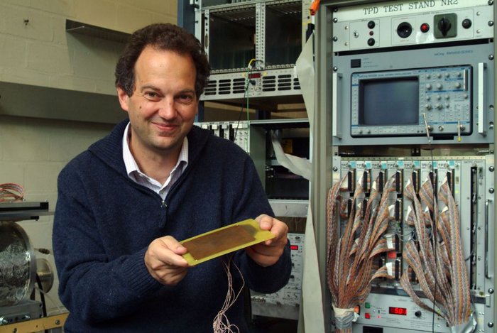 Achim Stahl von der RWTH Aachen Ein Mann mit krausem, kurzem Haar sitzt lächelnd neben einem Geräteschrank und zeigt eine Platine, die er in den Händen hält.