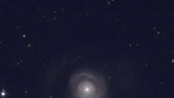 Im Zentrum des Bildes befindet sich eine difuse weißliche Wolke mit einem punktförmigen Leuchtenden Kern. Der HIntergrund ist blau-schwarz mit einzelnen weißes Punkten.