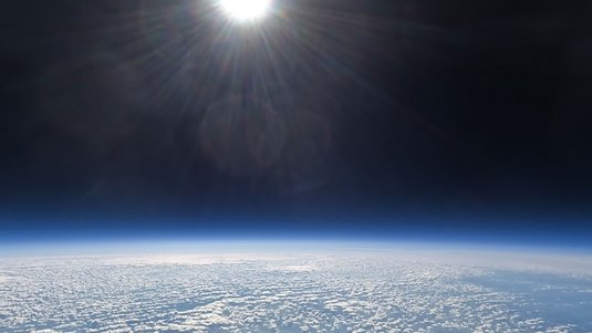 Blick aus der Erdatmopshäre: Am unteren Rand des Bildes ist die Erdoberfläche zu sehen, am oberen Rand eine angeschnittene Sonne - dazwischen ist das dunkle All.