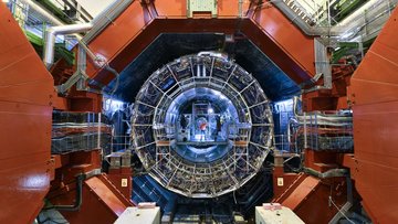 In einer mehreckigen, runden Öffnung befindet sich eine kreisrunde Röhre, die aus Metallgestänge besteht und im Inneren blau leuchtet; an ihrem anderen Ende steht ein Mensch.