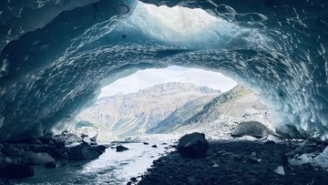 Blick aus einer Eishöhle, durch die ein kleiner Fluss fließt in eine alpine Landschaft